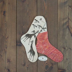 May include: Two wooden sock blockers featuring giraffe silhouettes, with the text "Mygidion" on one blocker. A pair of pink and orange knitted socks are displayed on the blockers. A small white tag with the text "Made with Mygidion" is on the table.