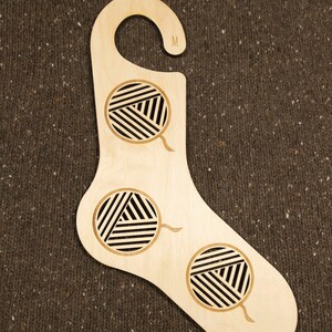 May include: A wooden sock-shaped hanger with a hook at the top. The hanger has three cutouts in the shape of balls of yarn. The hanger is labelled "M" on the hook.