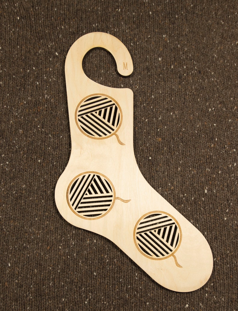 May include: A wooden sock-shaped hanger with a hook at the top. The hanger has three cutouts in the shape of balls of yarn. The hanger is labelled "M" on the hook.