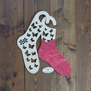 May include: A pair of pink knitted socks on a wooden sock blocker with a butterfly design. The sock blocker has a tag that says "Made with love, Mlyniedrow".