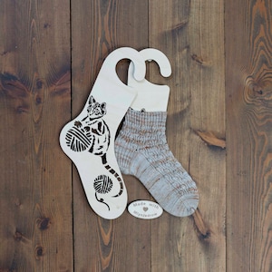 Op de afbeelding: Een houten sokken droogrek in de vorm van een kat die garenbollen vasthoudt. Het rek is wit met een zwart kat silhouet. Een paar beige en bruine sokken hangen aan het rek. Het rek heeft een klein label met de tekst "Made with ♥  Mynnjaddrow".