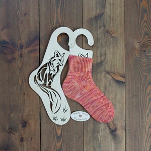 Op de afbeelding: Twee houten sokkenhangers met een vos-ontwerp. De hangers zijn wit en hebben een haak aan de bovenkant. Een paar roze en oranje sokken worden op een van de hangers getoond. De hangers hebben de tekst "Made with ♥ Mynjedrow" erop.