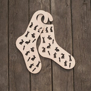 Könnte beinhalten: Zwei hölzerne Sockenhalter in Form von Socken mit einem Hasenmuster. Die Halter sind aus hellbraunem Holz und haben einen Haken oben.