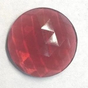 May include: A circular, faceted gemstone in a deep, translucent rose-red hue. The surface features a geometric pattern of triangular cuts that reflect light, creating a multi-dimensional appearance across the round, polished stone face.