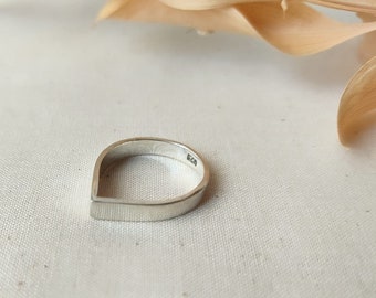 Op de afbeelding: Een zilveren ring met een druppelontwerp. De ring heeft een platte, rechthoekige basis die overgaat in een afgeronde bovenkant. De ring staat tegen een witte stoffen achtergrond met een gedroogd blad in de rechterbovenhoek.