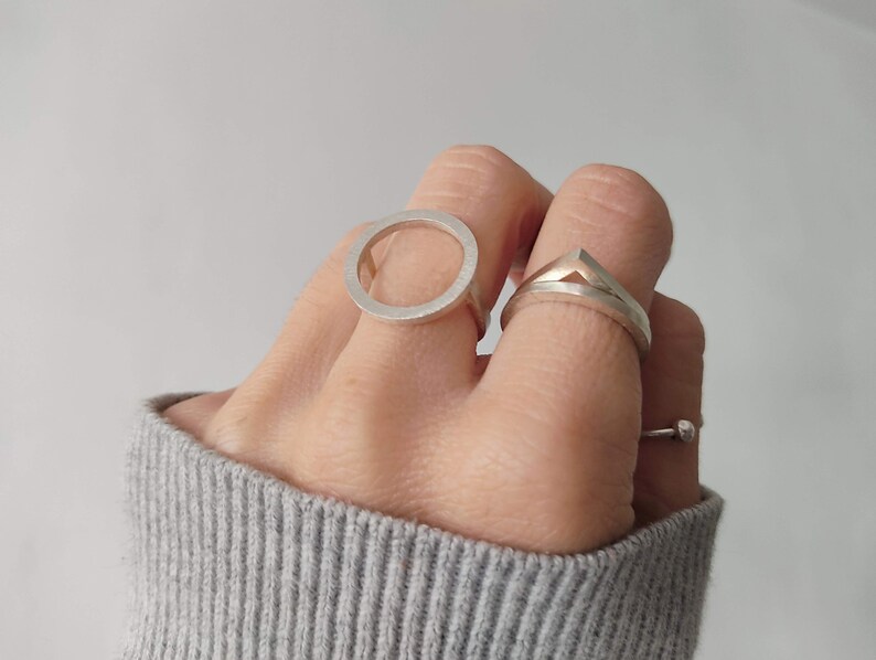 May include: Two silver rings on a hand. One ring is a simple circle, the other is a geometric shape with a pointed top.