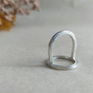 May include: A silver ring with a unique arch design. The ring is made of a smooth, polished metal and has a simple, modern aesthetic.