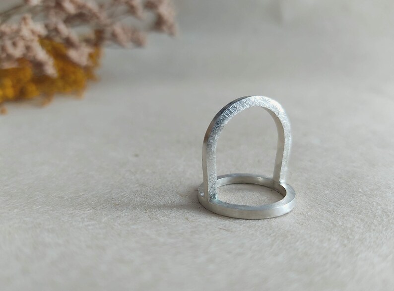 May include: A silver ring with a unique arch design. The ring is made of a smooth, polished metal and has a simple, modern aesthetic.