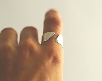 May include: A silver ring with a geometric design. The ring features a split design with a triangular cutout, creating a modern and minimalist aesthetic. The ring is worn on a finger, showcasing its sleek and polished surface.