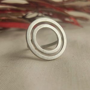 May include: A silver ring with a double circle design. The ring is made of a smooth, brushed metal.