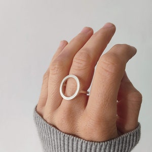 May include: A silver ring with a large, open circle design. The ring is worn on the middle finger of a hand.