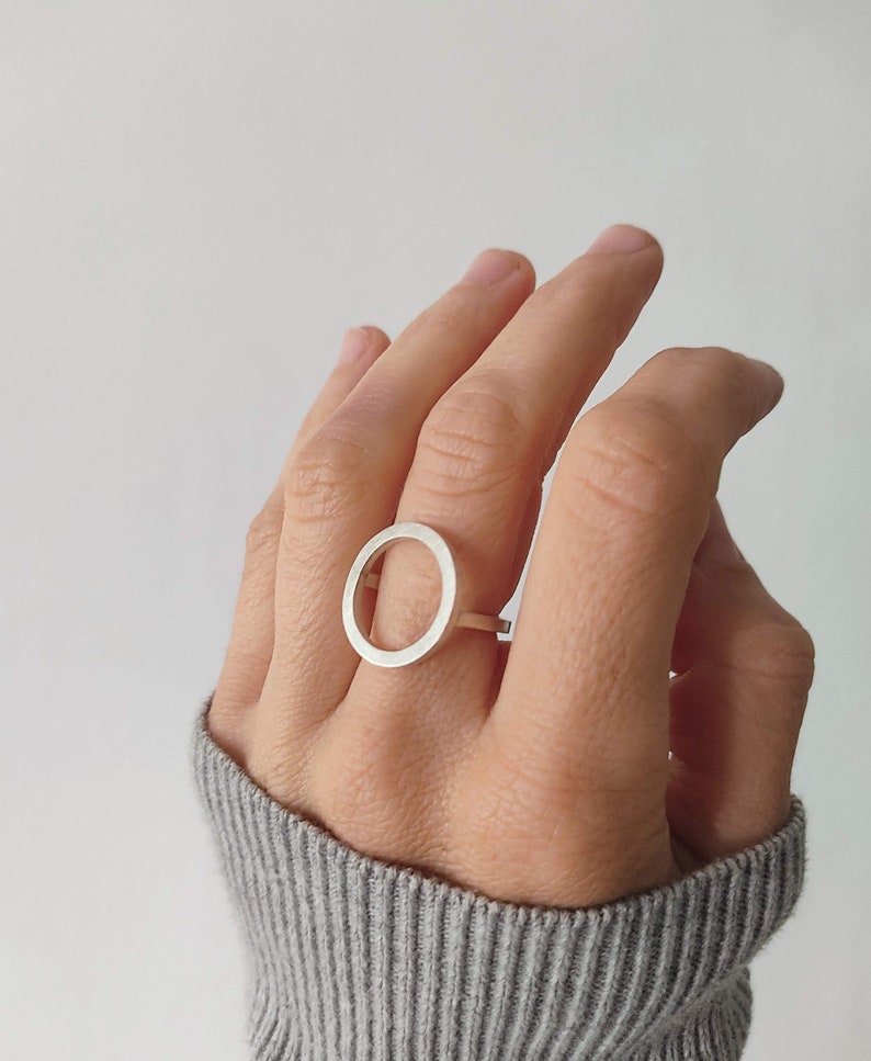 May include: A silver ring with a large, open circle design. The ring is worn on the middle finger of a hand.