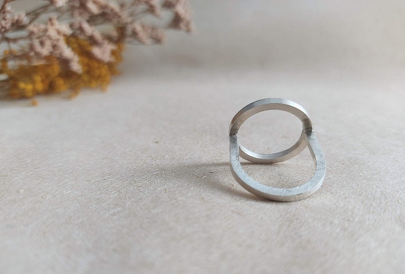 May include: A silver ring with a unique design featuring two interlocking circles. The ring is made of polished metal and has a smooth, modern look.