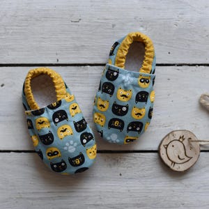 Peut inclure: Une paire de chaussons bleus pour b&eacute;b&eacute; avec une bordure jaune et un motif de chats noirs et jaunes.