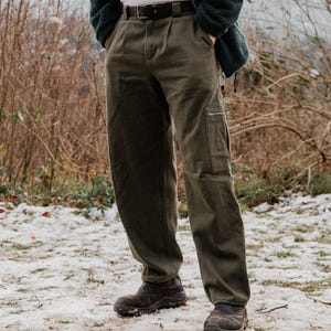 May include: Olive green cargo pants with a side pocket and a zipper. The pants have a pleated front and are worn with a brown belt. The person is wearing dark brown shoes. The background is a snowy, outdoor scene.
