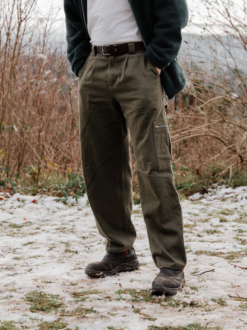 May include: Olive green cargo pants with a side pocket and a zipper. The pants have a pleated front and are worn with a brown belt. The person is wearing dark brown shoes. The background is a snowy, outdoor scene.