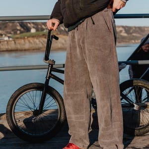 May include: A person wearing gray pants with red stitching and red shoes stands next to a black BMX bike. The person is wearing a black jacket. The background includes a body of water and a bridge.