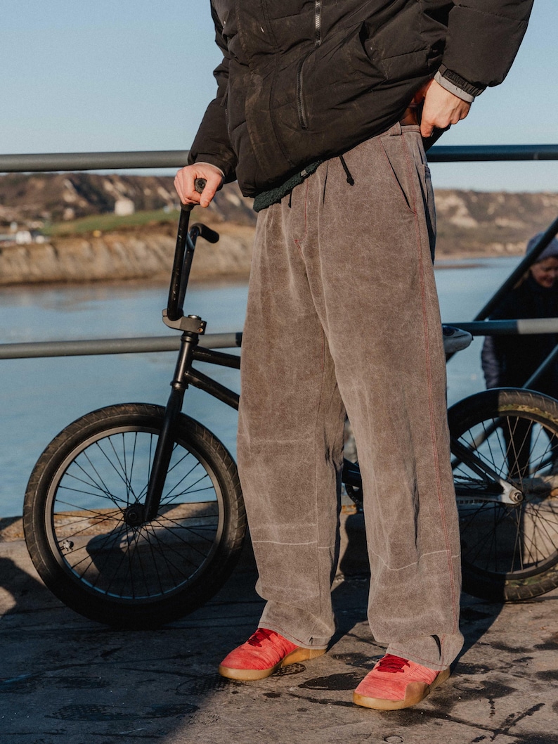 May include: A person wearing gray pants with red stitching and red shoes stands next to a black BMX bike. The person is wearing a black jacket. The background includes a body of water and a bridge.