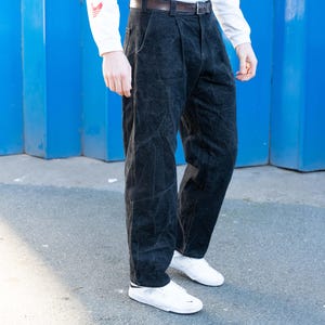 May include: Black corduroy pants with a pleated front, paired with a white long-sleeve shirt and white sneakers. The pants have a dark brown belt. The background is a bright blue wall.