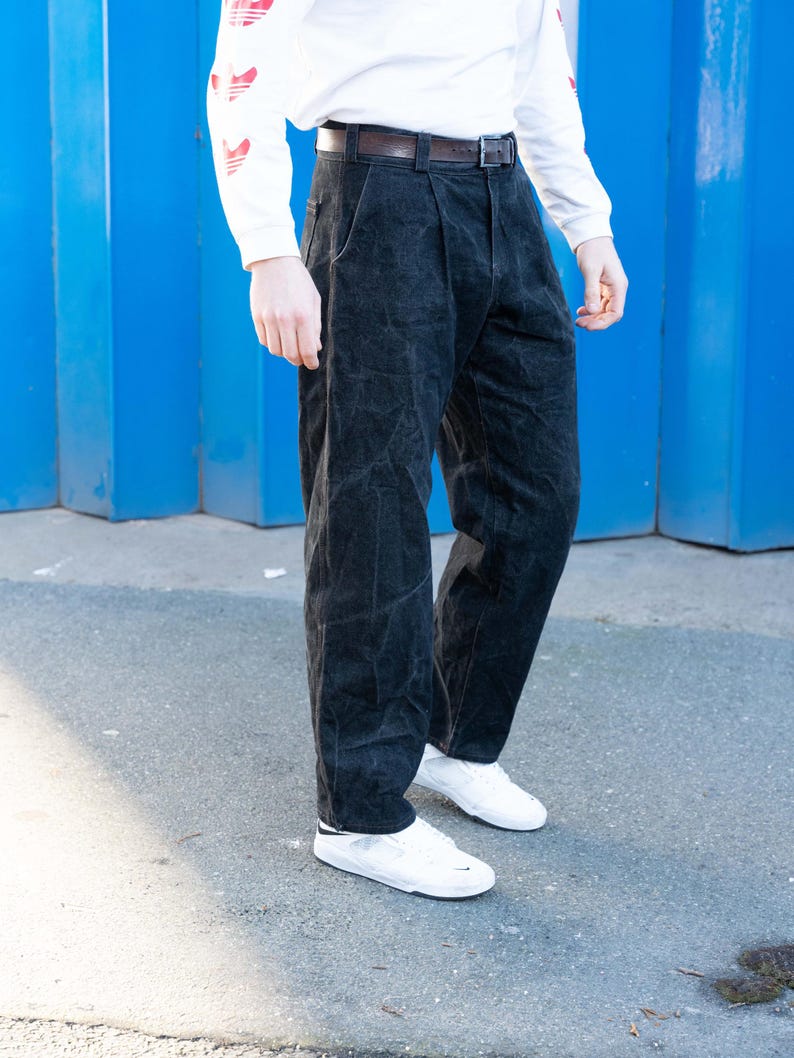 May include: Black corduroy pants with a pleated front, paired with a white long-sleeve shirt and white sneakers. The pants have a dark brown belt. The background is a bright blue wall.