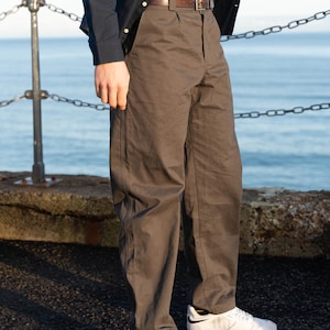 May include: A person wearing a dark navy button-down shirt, a black t-shirt, and brown pants with a brown belt. The person is also wearing white sneakers with black accents. The background features a body of water and a chain railing.