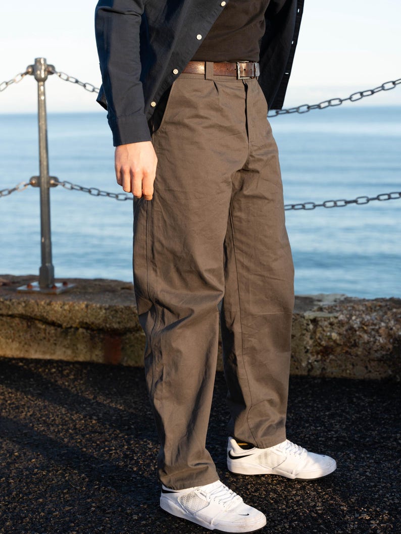 May include: A person wearing a dark navy button-down shirt, a black t-shirt, and brown pants with a brown belt. The person is also wearing white sneakers with black accents. The background features a body of water and a chain railing.