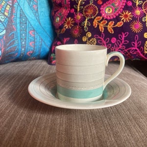 May include: A white and gray striped coffee cup with a turquoise band and a matching saucer. The cup has a handle and is sitting on a brown couch.