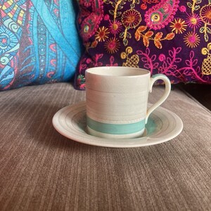 Puede incluir: Una taza de café de cerámica blanca con una franja azul y un platillo a juego. La taza y el platillo están sobre un sofá marrón.