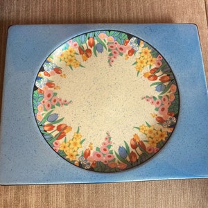 Könnte beinhalten: Ein blauer quadratischer Teller aus Keramik mit einer weißen Mitte und einem floralen Design am Rand. Die Blumen sind in den Farben Rosa, Gelb und Blau gehalten.