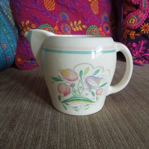 Könnte beinhalten: Eine weiße Keramikkanne mit Blumenmuster. Die Kanne hat ein hellrosa, grünes und blaues Blumenmuster mit einem hellrosa und grünen Rand am oberen Rand.