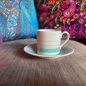 Puede incluir: Una taza de café de cerámica blanca con una franja turquesa y un platillo. La taza y el platillo están sobre un sofá marrón con almohadas coloridas.