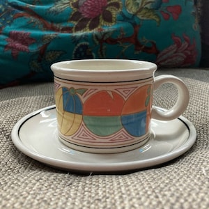 Könnte beinhalten: Eine weiße Teetasse und Untertasse aus Keramik mit einem farbenfrohen geometrischen Design. Die Tasse hat einen Henkel und die Untertasse einen schwarzen Rand.