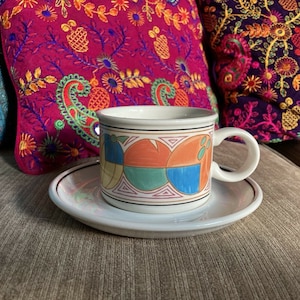 May include: A white ceramic teacup and saucer with a colourful geometric design. The cup has a rounded handle and the saucer has a gold rim.