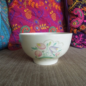 Könnte beinhalten: Eine weiße Keramikschüssel mit einem Blumendesign mit rosa, lila und grünen Blumen. Die Schüssel steht auf einem braunen Sofa mit bunten Kissen.