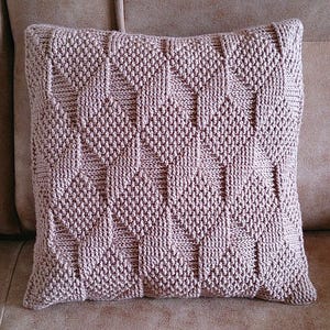 May include: A square, knitted pillow in a muted mauve color. The pillow features a geometric pattern of raised diamonds and textured stitches. It is placed on a brown sofa, showcasing its decorative design and cozy texture.