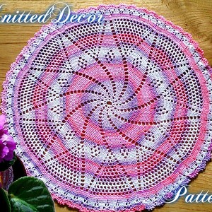 Puede incluir: Un tapete circular hecho a mano, con un dise&ntilde;o en espiral en tonos rosa, blanco y morado. El tapete est&aacute; tejido a ganchillo con un delicado patr&oacute;n calado y tiene un borde festoneado. Las palabras "Knitted Decor" y "Pattern" est&aacute;n escritas en la imagen.