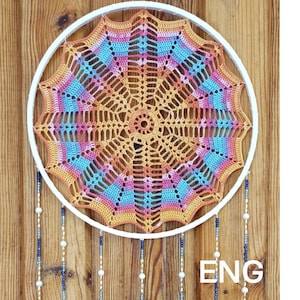May include: A circular white hoop containing a multi-colored crocheted mandala in shades of orange, pink, and blue. Hanging from the bottom are several vertical strands decorated with small white beads and dark blue seed beads. The text ENG is visible.