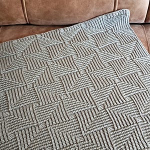 May include: A light gray knitted blanket with a geometric pattern of interwoven lines. The blanket is rectangular and has a textured surface. The blanket is folded over a brown leather couch.