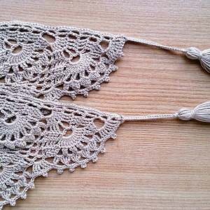 May include: Close-up of a beige crocheted lace trim with a floral design. The trim has two tassels at the ends. The lace is laid on a wooden surface. This handmade trim is perfect for craft projects.
