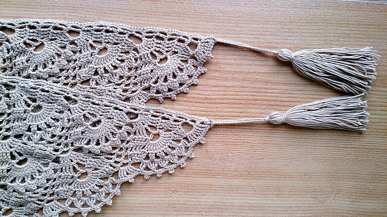 May include: Close-up of a beige crocheted lace trim with a floral design. The trim has two tassels at the ends. The lace is laid on a wooden surface. This handmade trim is perfect for craft projects.