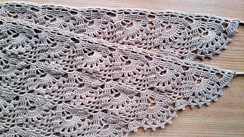 May include: Two pieces of beige crocheted lace with a delicate floral pattern. The lace has a scalloped edge and is displayed on a wooden surface. This handmade lace is suitable for various craft projects.