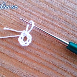 Pu&ograve; includere: Primo piano di un progetto all'uncinetto in corso. Un filo bianco &egrave; avvolto attorno a un uncinetto argentato con un manico verde, formando un piccolo cerchio. Il testo "Knitted Decor" &egrave; nell'angolo in alto a sinistra.