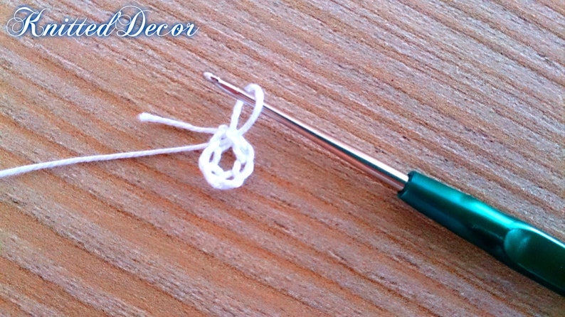 Pu&ograve; includere: Primo piano di un progetto all'uncinetto in corso. Un filo bianco &egrave; avvolto attorno a un uncinetto argentato con un manico verde, formando un piccolo cerchio. Il testo "Knitted Decor" &egrave; nell'angolo in alto a sinistra.