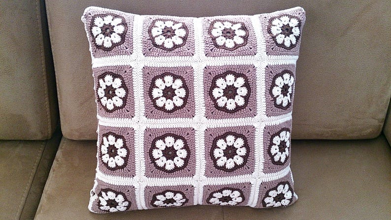 May include: A crocheted pillow with a brown and white floral pattern. The pillow is made of a soft, textured yarn and has a square shape.