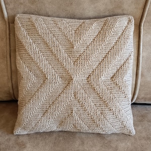 Könnte beinhalten: Ein quadratisches, beiges Zierkissen mit einem strukturierten, geometrischen Muster. Das Kissen liegt auf einem beigen Sofa. Das Design zeigt eine zentrale Kreuzform mit diagonalen Linien, die nach außen strahlen und ein strukturiertes Aussehen erzeugen.