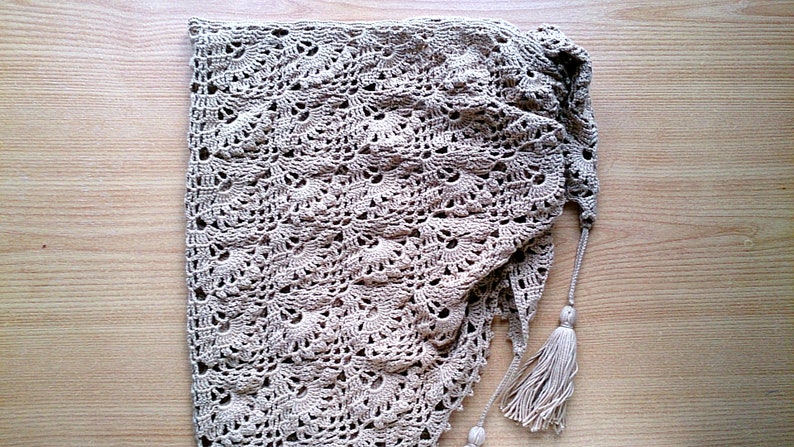 May include: A beige, handmade crochet shawl with a delicate, openwork pattern. The shawl is folded, revealing its intricate design and texture. Tassels hang from the edge, adding a decorative element to this cozy accessory.