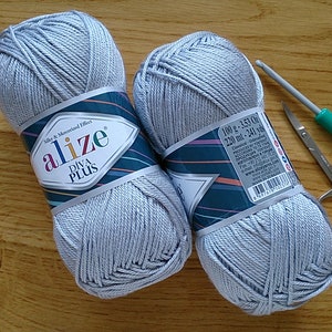 May include: Two grey yarn skeins, a crochet hook, small scissors, and clear buttons on a wooden surface. The yarn is labelled "Alize Diva Plus". The crochet hook is green and silver. The buttons are transparent.