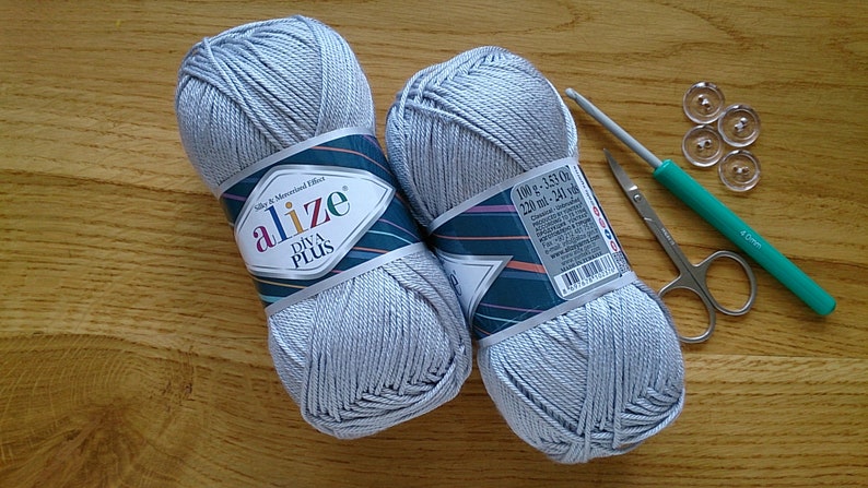 May include: Two grey yarn skeins, a crochet hook, small scissors, and clear buttons on a wooden surface. The yarn is labelled "Alize Diva Plus". The crochet hook is green and silver. The buttons are transparent.