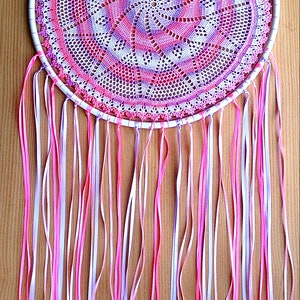 Puede incluir: Un atrapasue&ntilde;os hecho a mano con un marco circular blanco. El centro presenta un doily de ganchillo detallado en tonos de rosa, morado y blanco. Largas cintas que fluyen en rosa, blanco y lavanda cuelgan de la parte inferior.
