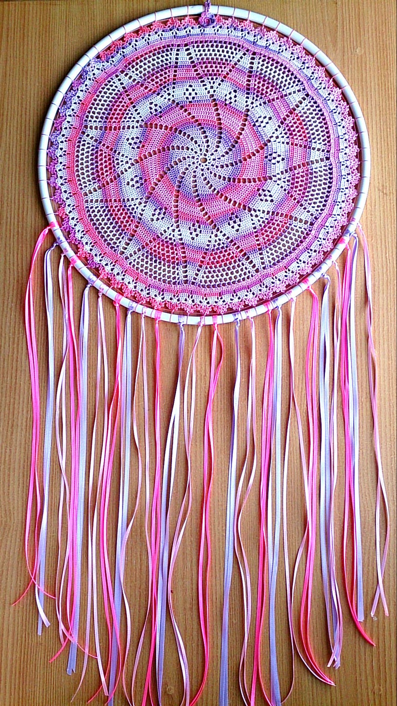 Puede incluir: Un atrapasue&ntilde;os hecho a mano con un marco circular blanco. El centro presenta un doily de ganchillo detallado en tonos de rosa, morado y blanco. Largas cintas que fluyen en rosa, blanco y lavanda cuelgan de la parte inferior.